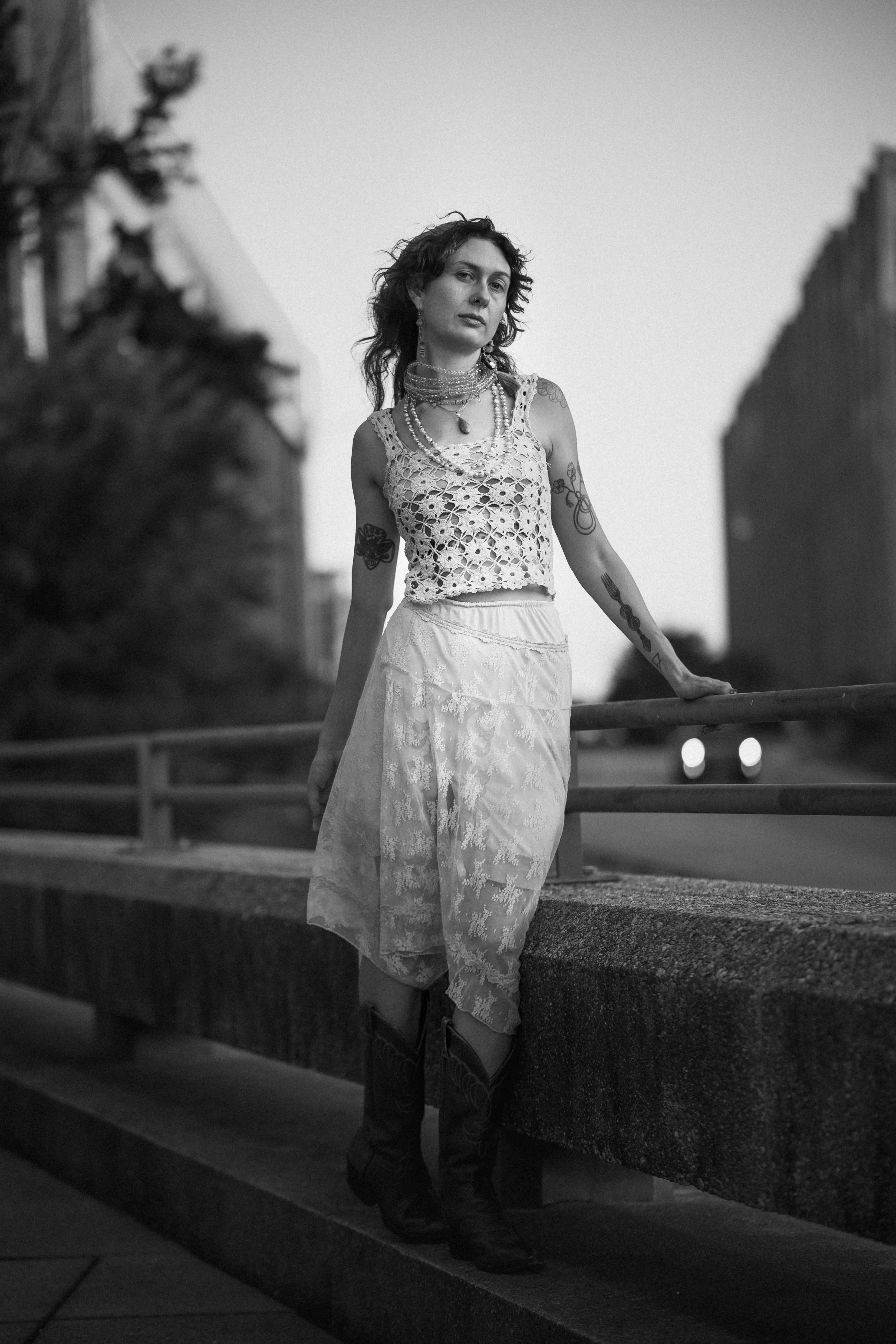 A woman in a crocheted top, layered necklaces, and cowboy boots leans against a bridge railing at dusk, city buildings and passing headlights softly blurred behind her, in black and white.