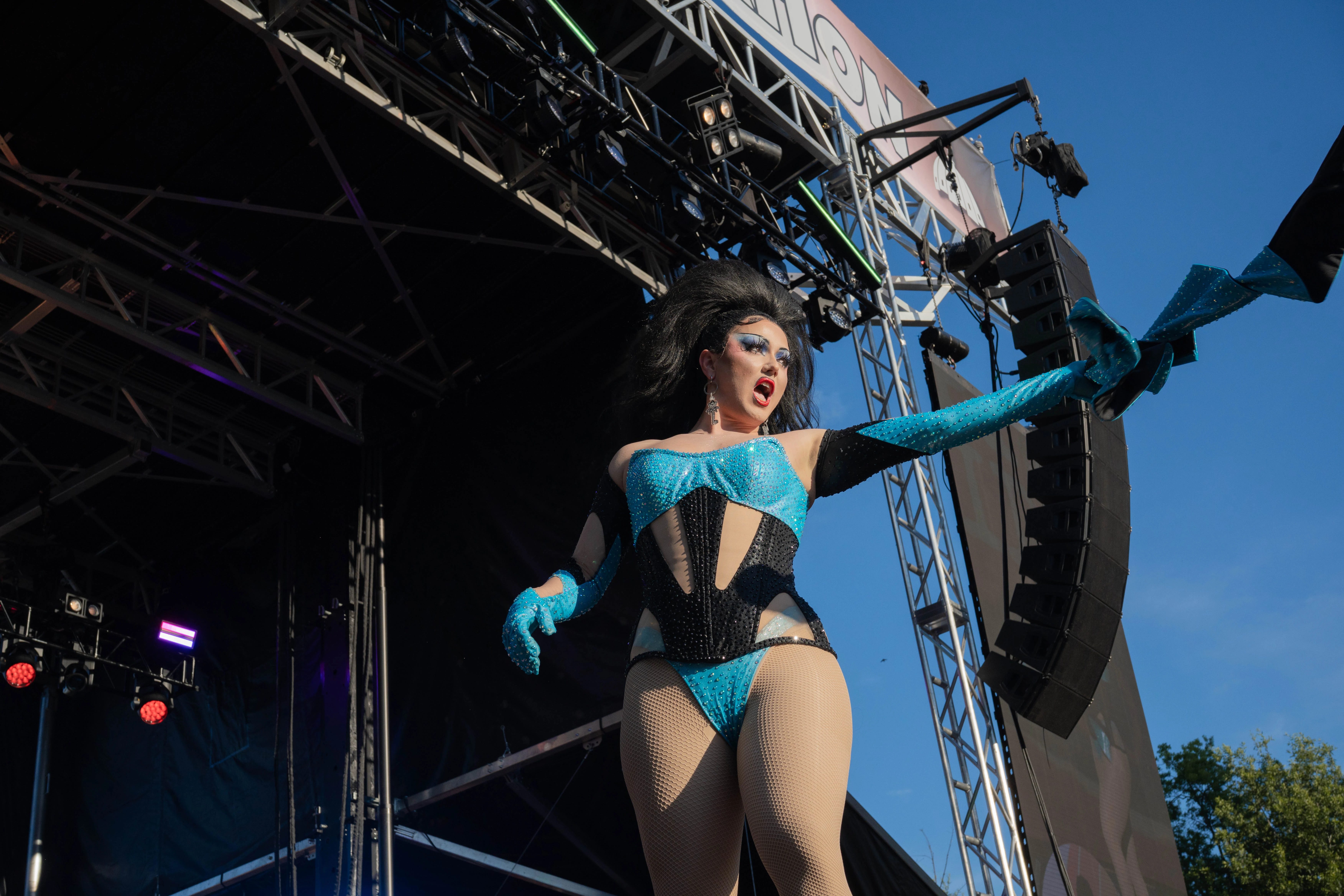 A drag performer in a teal costume throws both arms wide on an outdoor festival stage beneath a clear blue sky.