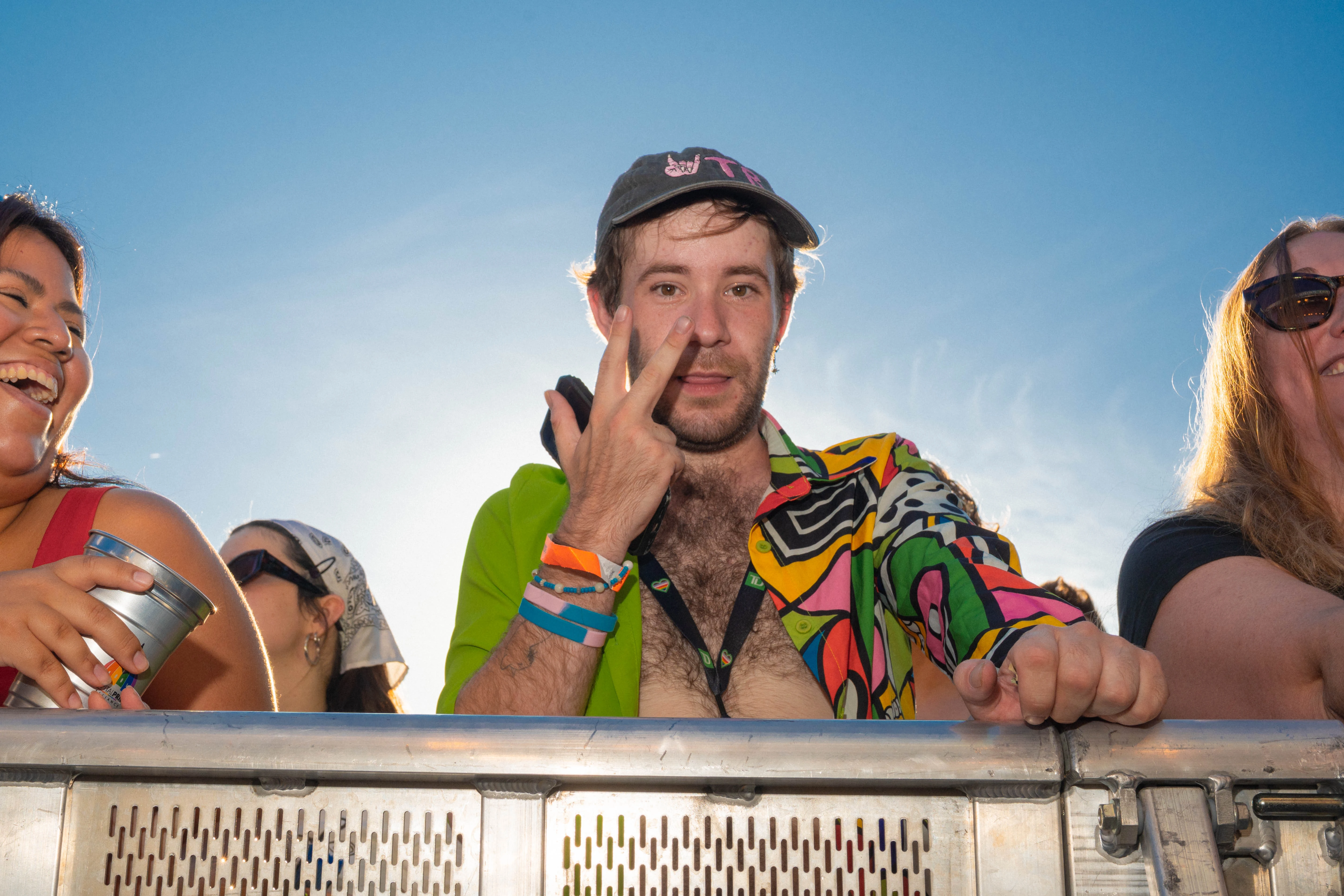 A festival-goer in a bright shirt leans on a metal barricade in the sun, flashing a peace sign, flanked by others.