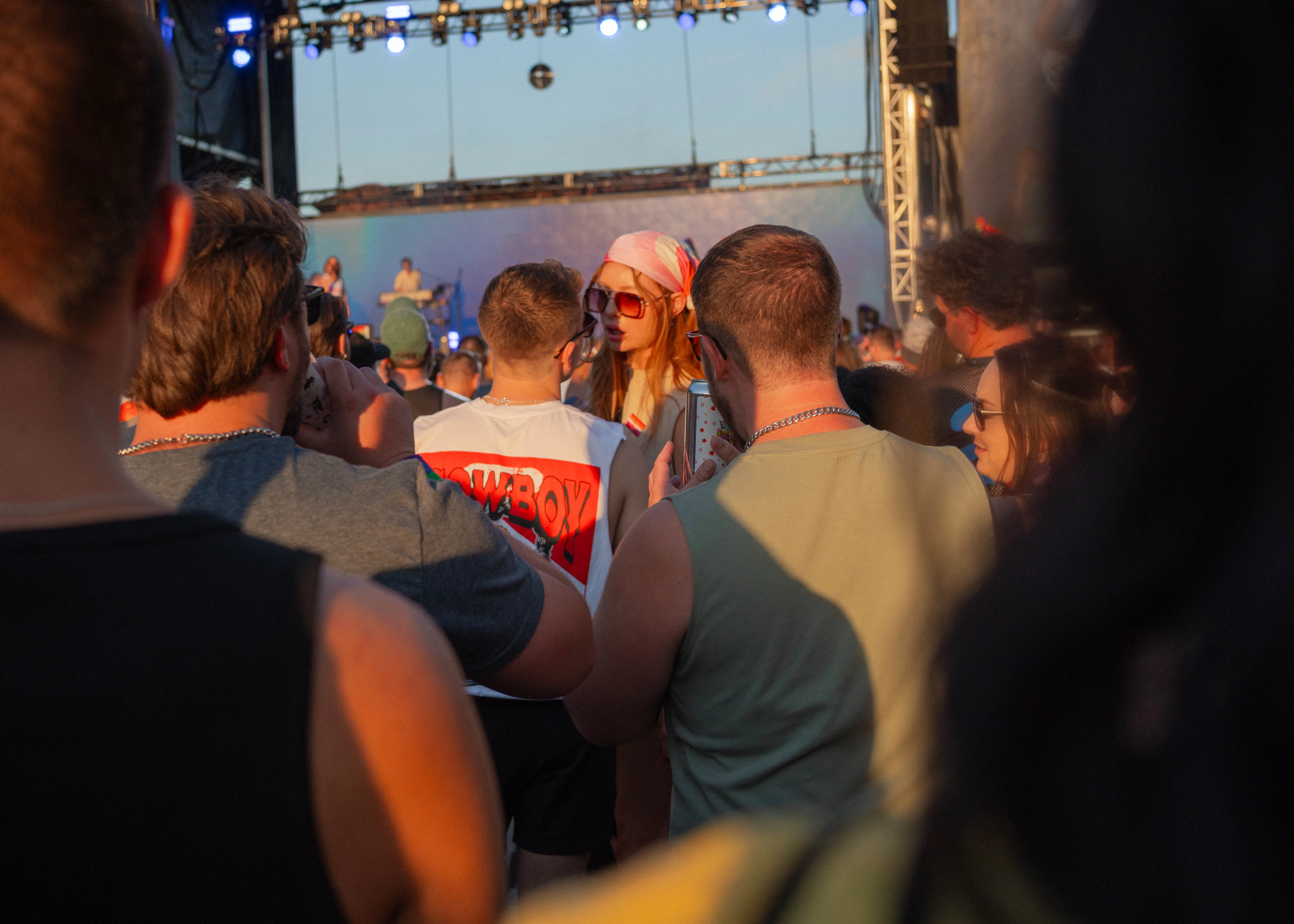 A concert crowd in warm low sun; a person in a red cap and sunglasses turns toward the camera as the stage glows behind.