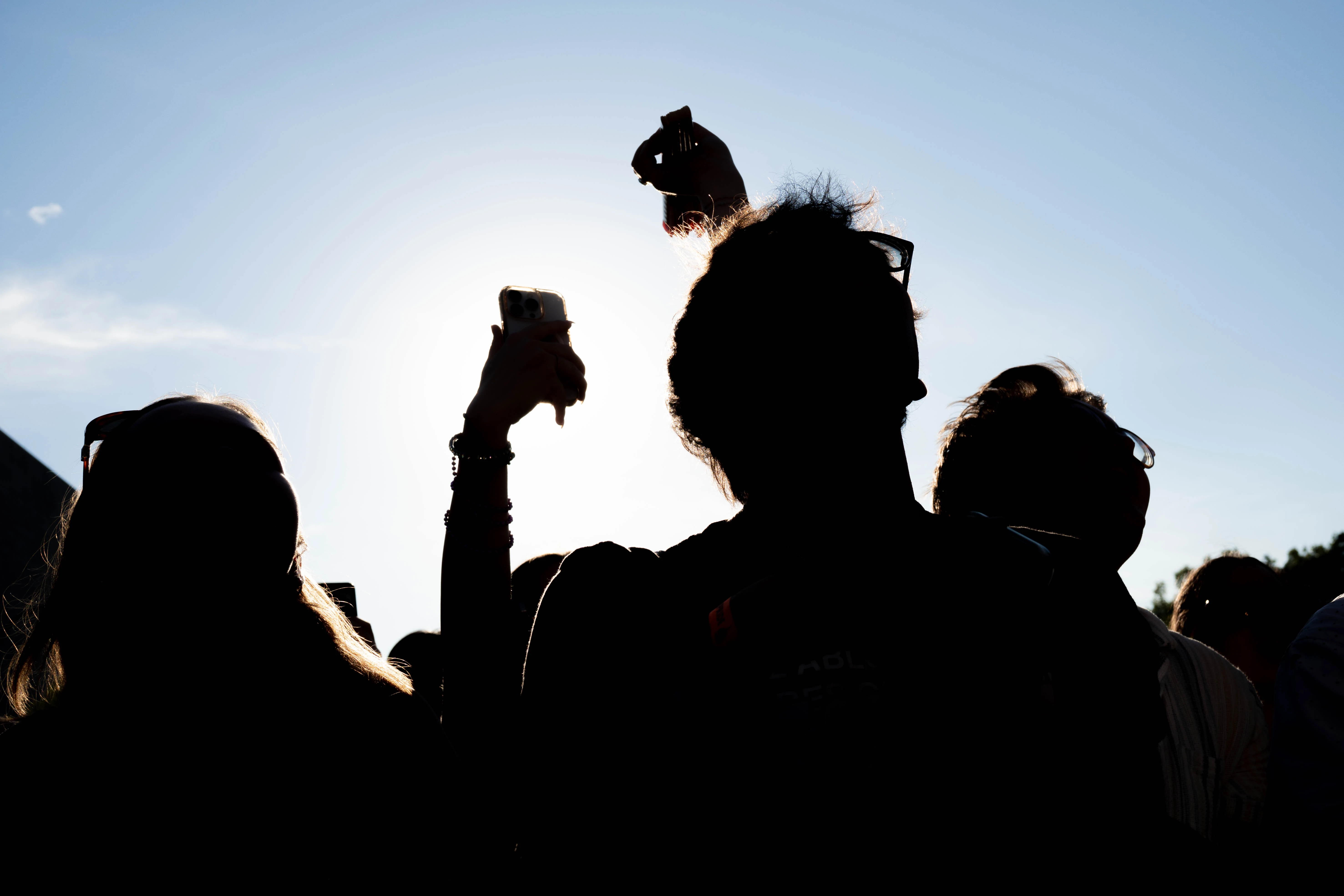 Backlit silhouettes of a crowd against a bright sky, hands and a phone raised overhead.