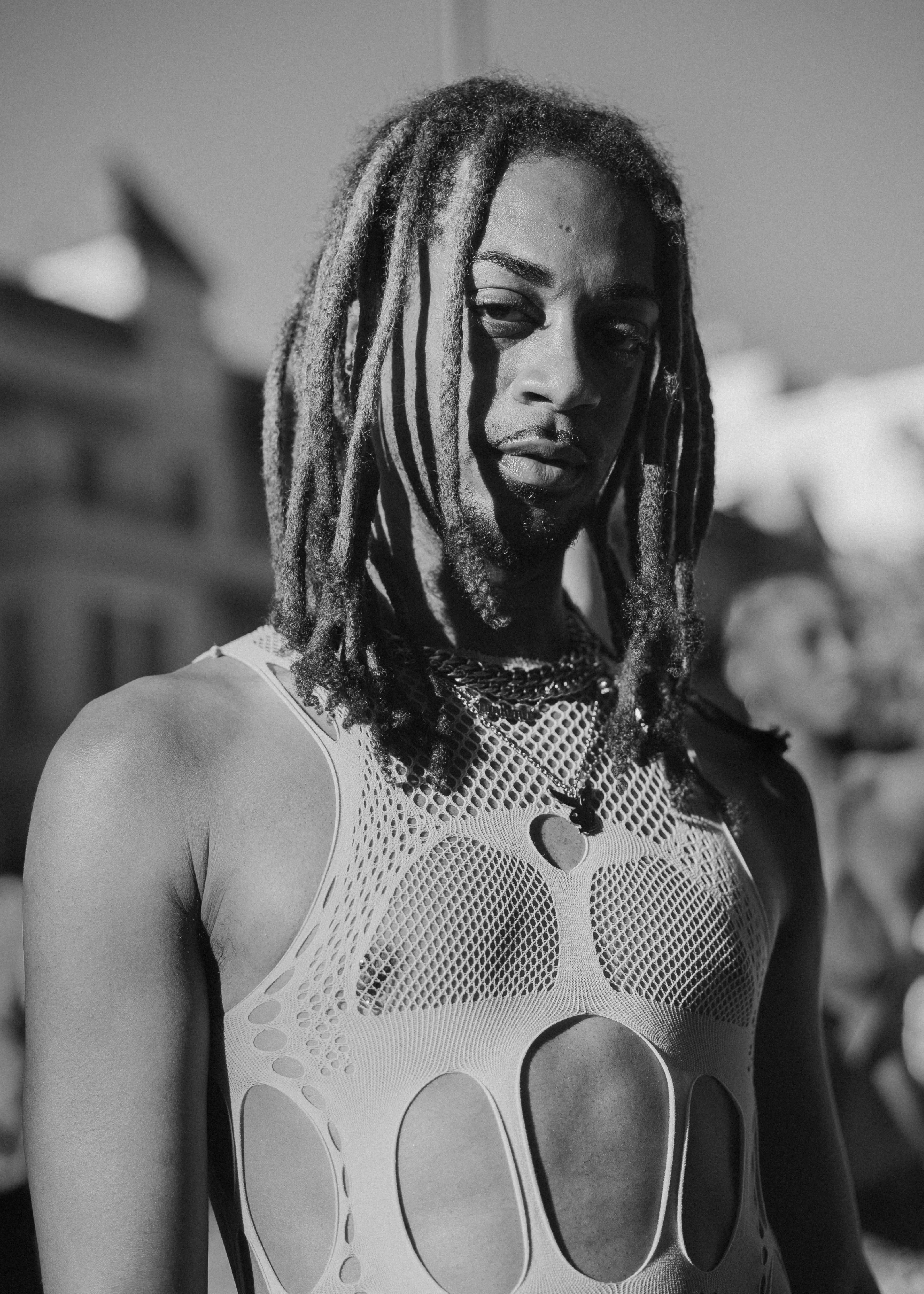 A close portrait of a person with long locs and layered chain necklaces, wearing a cut-out mesh top, lit in warm side light.
