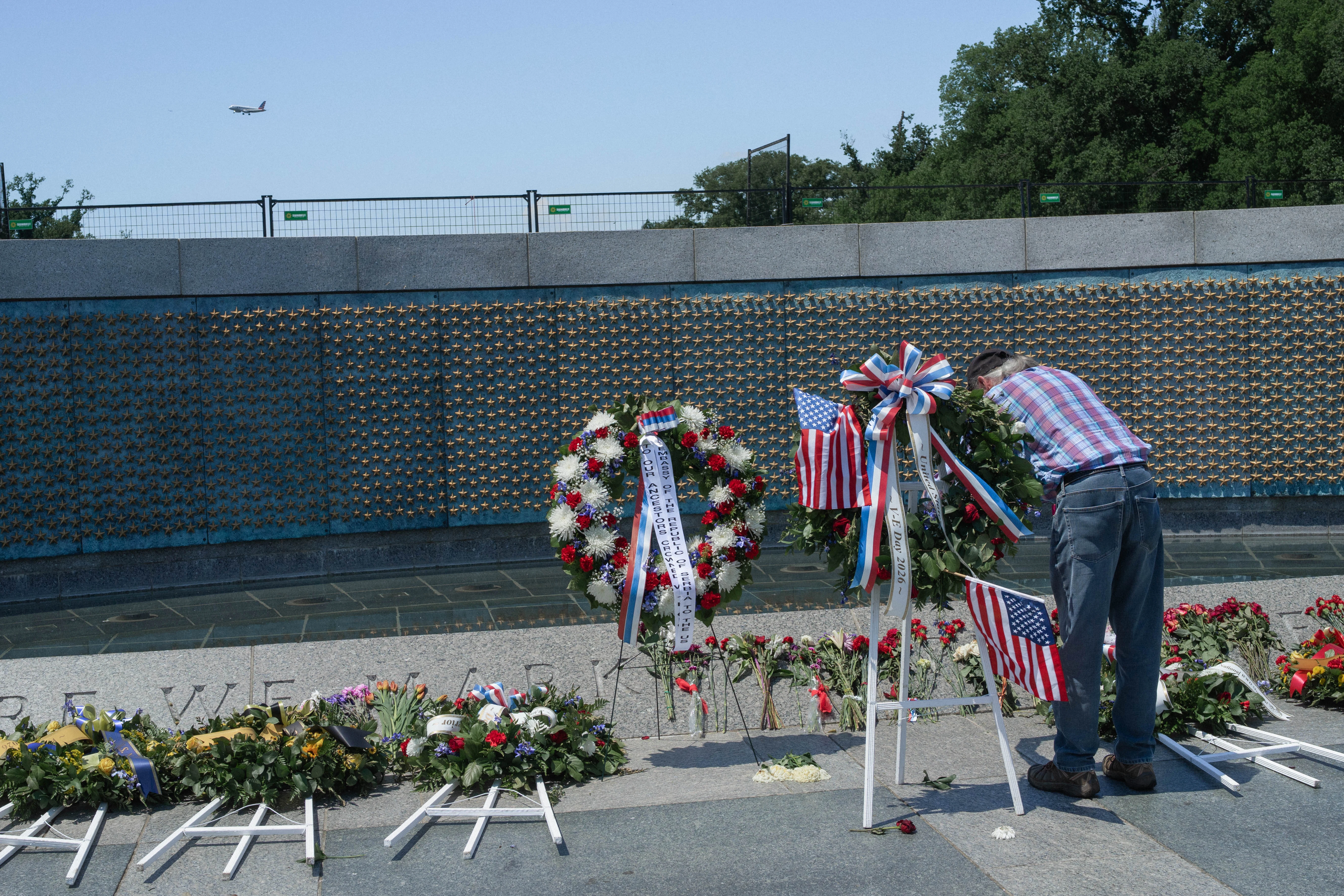 A man in a stars-and-stripes shirt arranges flags and wreaths at a memorial wall as a plane crosses the sky, Washington, D.C.