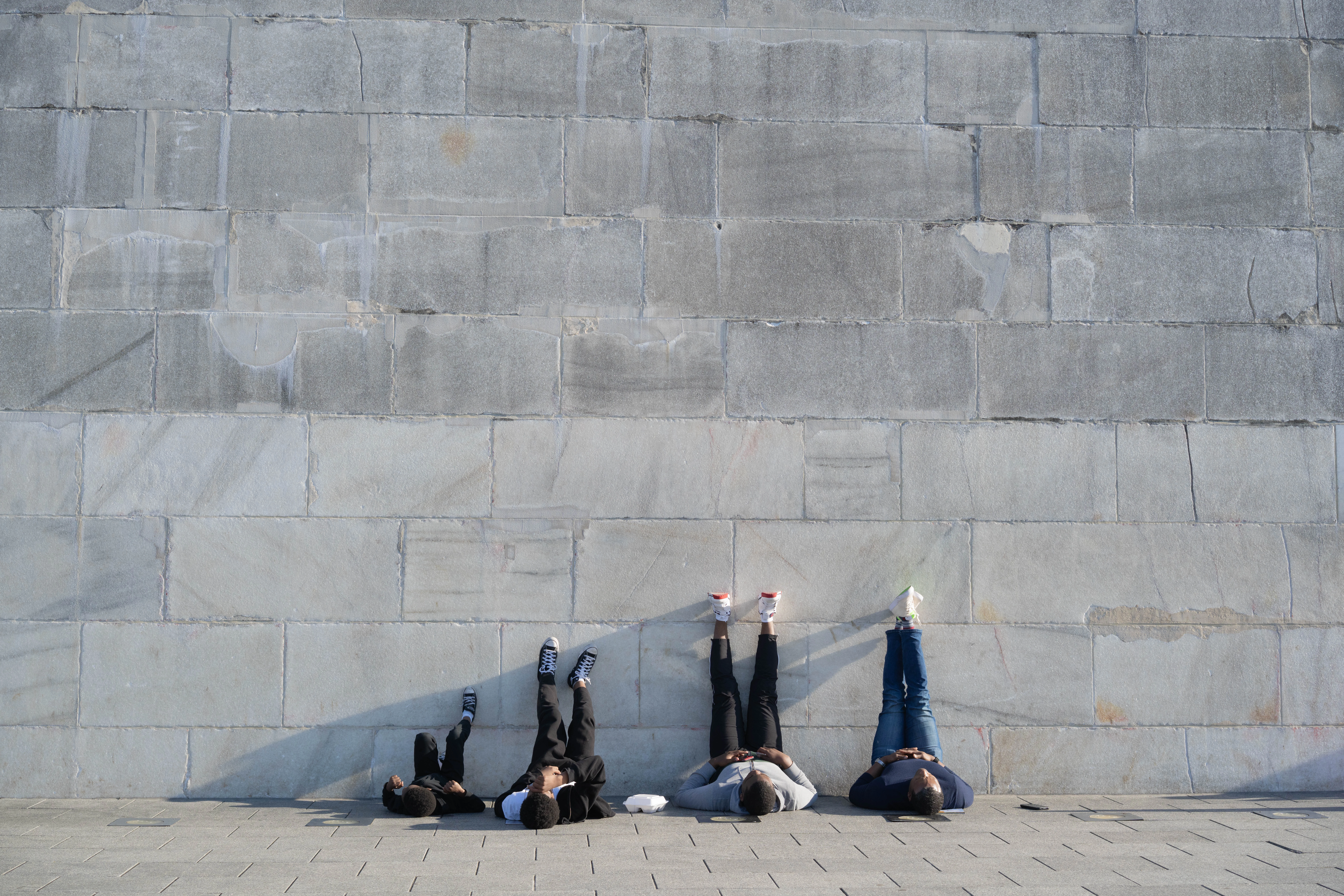 Several people lie on their backs along the base of a vast pale stone monument wall in hard sun, feet up against the stone, Washington, D.C.